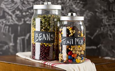 A large jar containing three kinds of dry beans with a chalkboard label that reads “Beans,” and a smaller jar filled with nuts, raisins and chocolate candy with a chalkboard label that reads “Trail Mix.” 